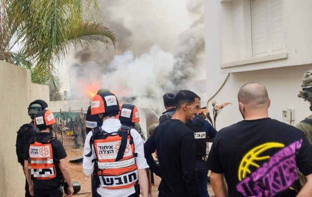 Several volunteers in black, orange and white gear with the United Hatzaleh service face smoke and fire after a rocket attack on the village.
